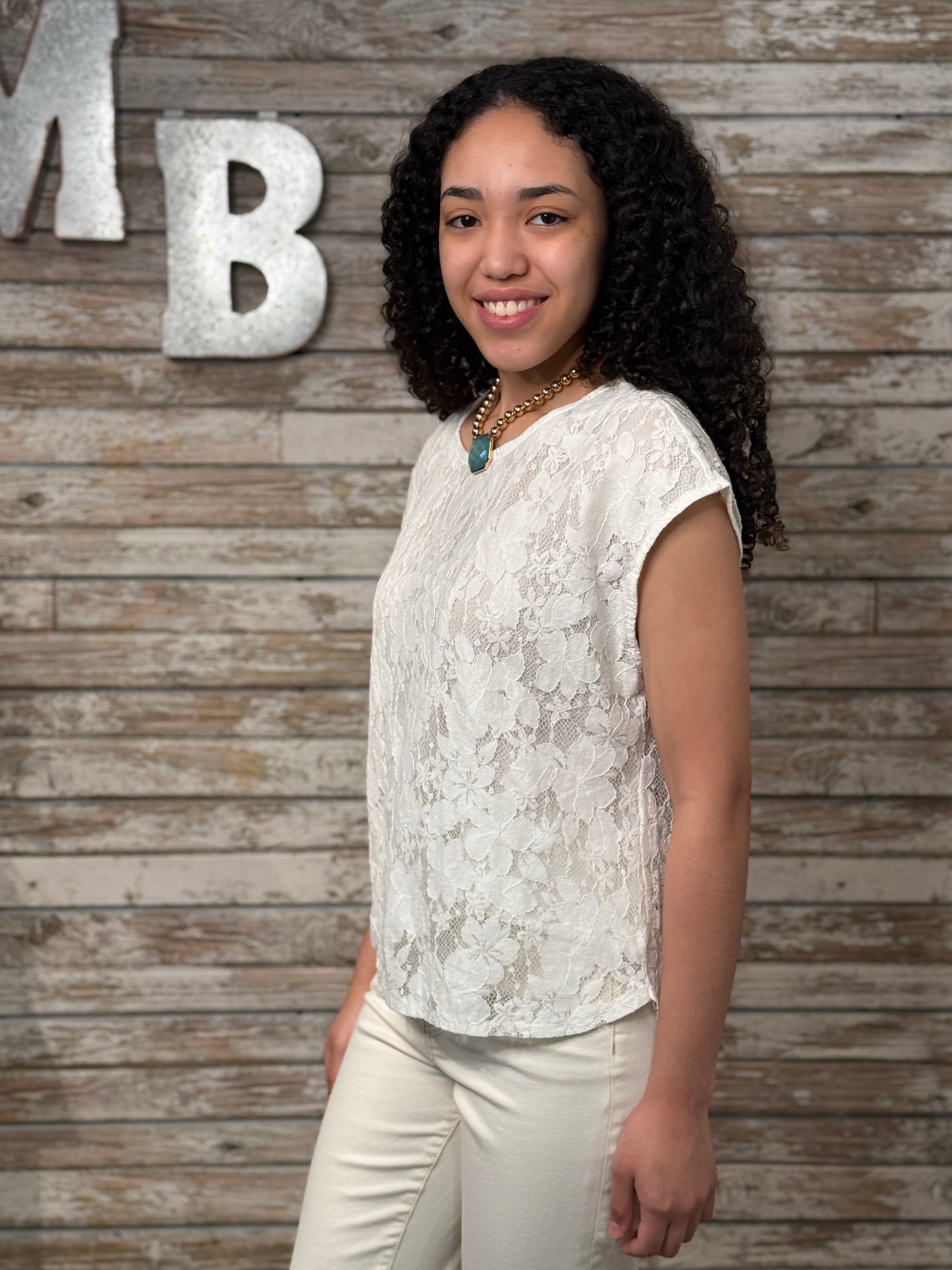 Woman in a white lace top and beige pants standing against a wooden wall with decorative letters.