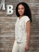 Woman in a white lace top and beige pants standing against a wooden wall with decorative letters.