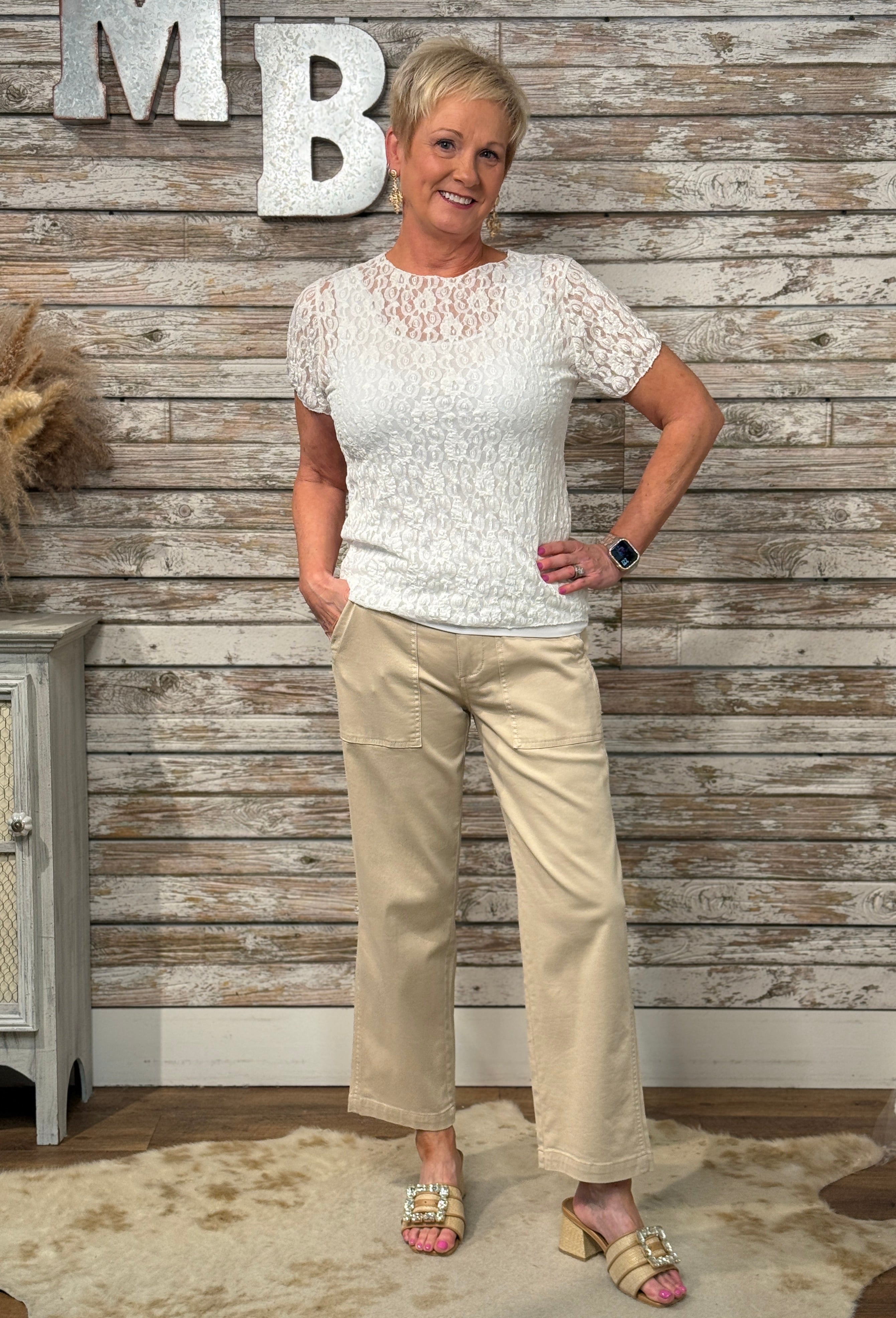 Woman in a white lace top and beige pants standing against a wooden wall with 'MB' letters.