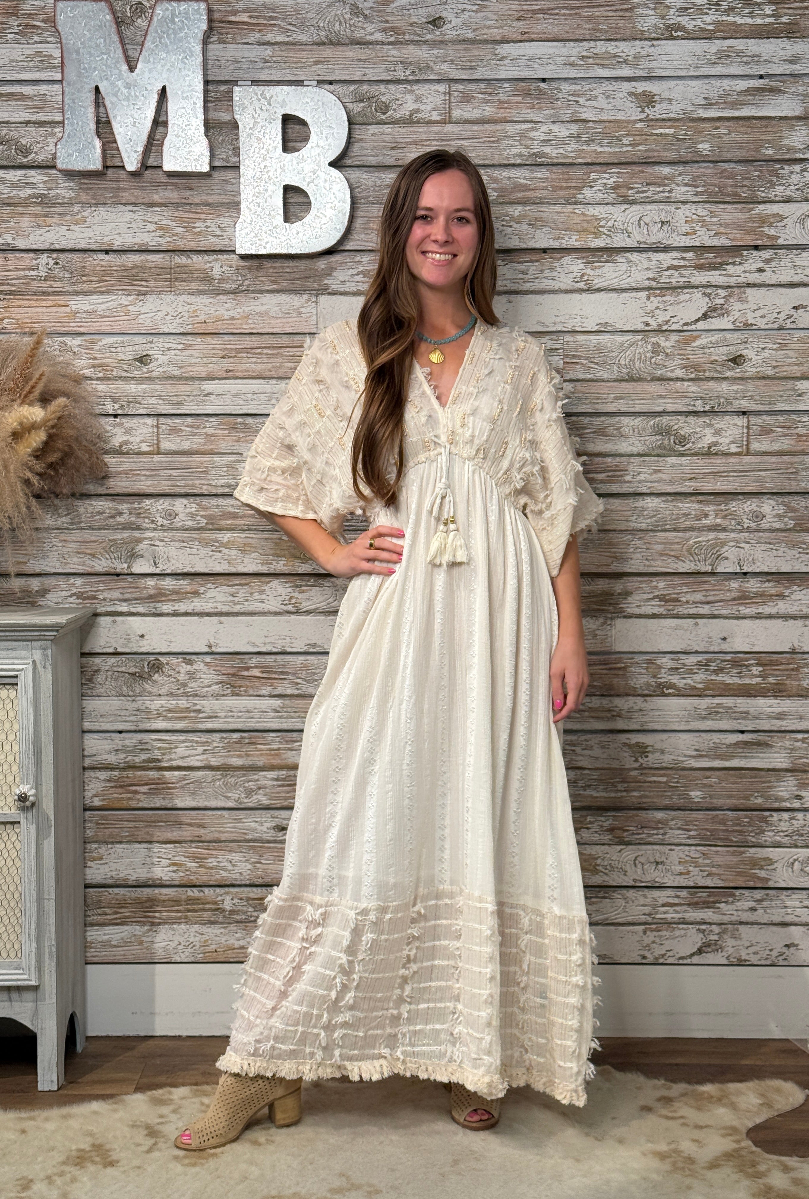 Woman wearing a cream boho maxi dress with embroidered detailing, tassel tie neckline, flowy short sleeves, textured fabric, and a relaxed tiered silhouette.