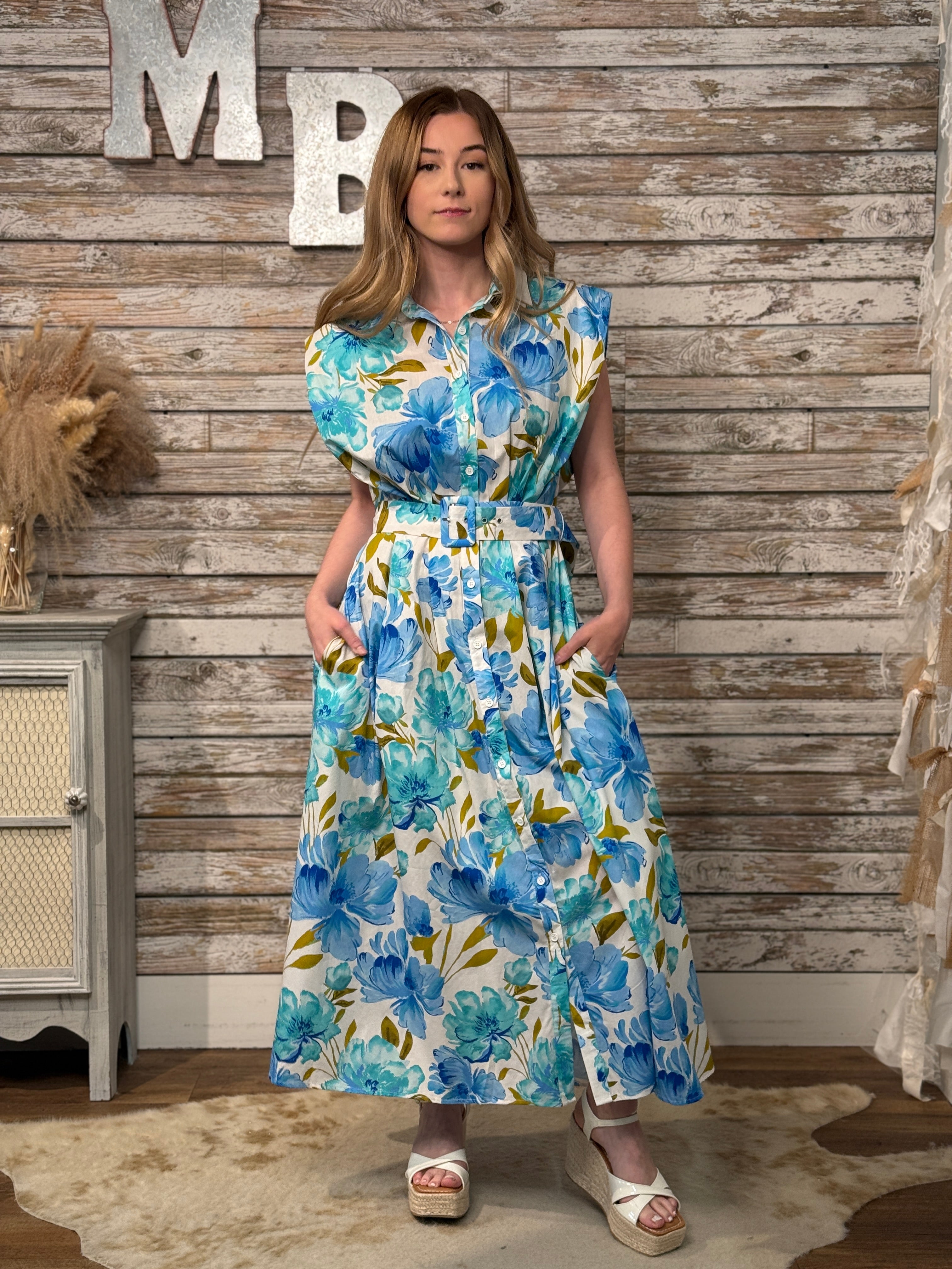 Woman wearing a sleeveless blue floral midi dress with structured shoulders, collared neckline, button-front detail, matching belt, side pockets, and an A-line silhouette.