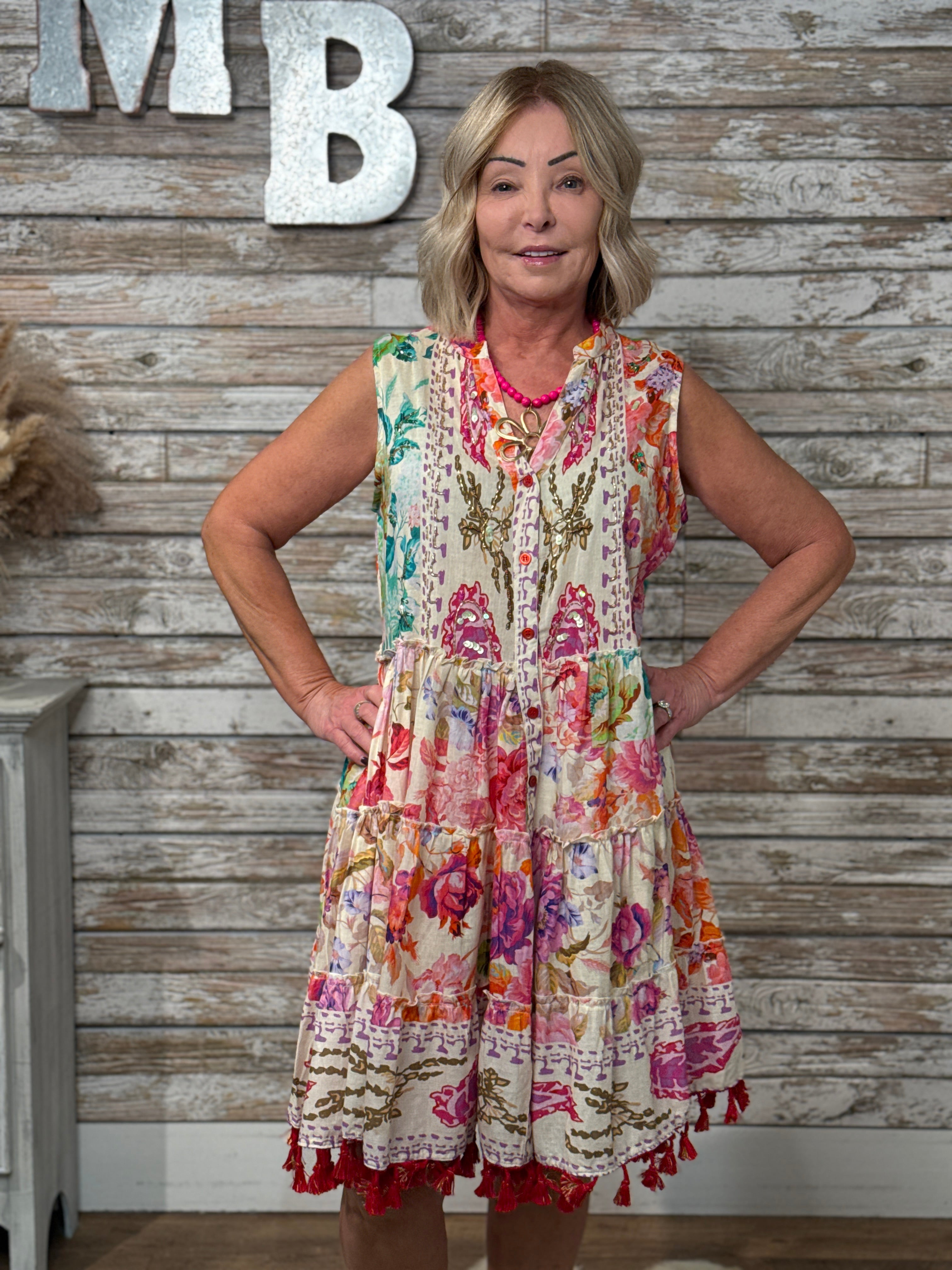 Woman wearing a sleeveless boho floral mini dress with tiered ruffle layers, mixed floral patchwork print, tassel hem detailing, and a relaxed flowy fit.