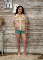Woman wearing a colorful striped shirt and green shorts standing against a wooden wall.