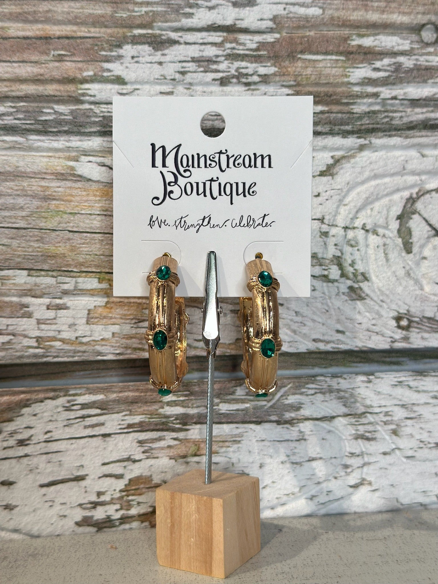 Gold hoop earrings with green stones on a wooden stand against a rustic wood background.
