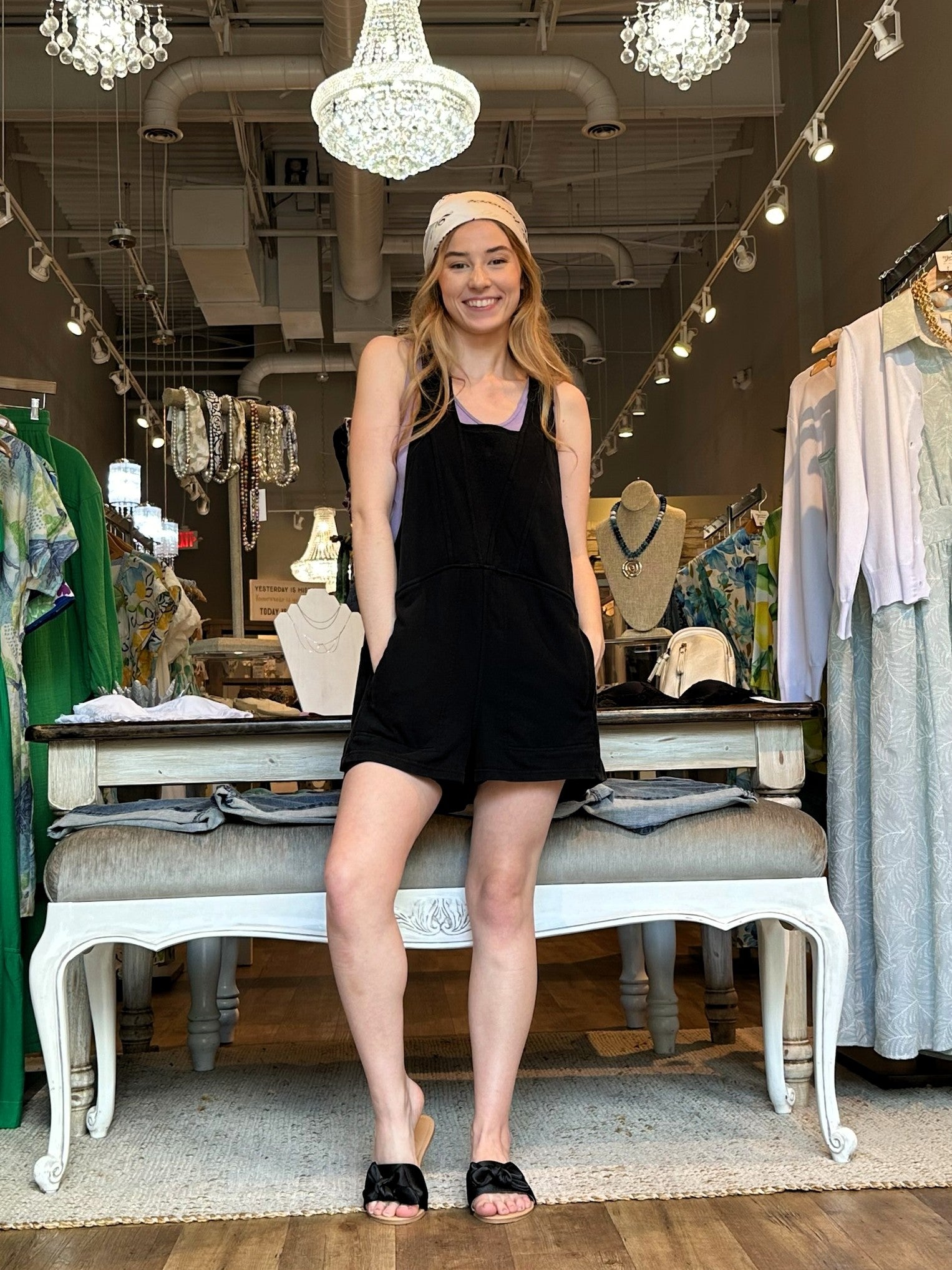 A woman modeling a black sleeveless romper with side pockets, styled with a beige headband and black slide sandals, standing on a white bench inside a boutique.