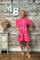 A woman modeling a bright pink short-sleeve dress with white trim, standing in front of a rustic whitewashed wood wall with large decorative letters 'MB' and a small cabinet to the side.