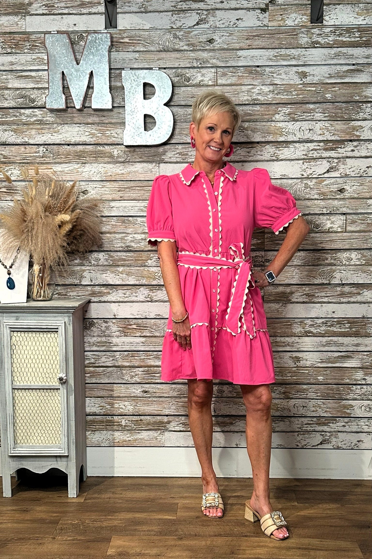 A woman modeling a bright pink short-sleeve dress with white trim, standing in front of a rustic whitewashed wood wall with large decorative letters 'MB' and a small cabinet to the side.