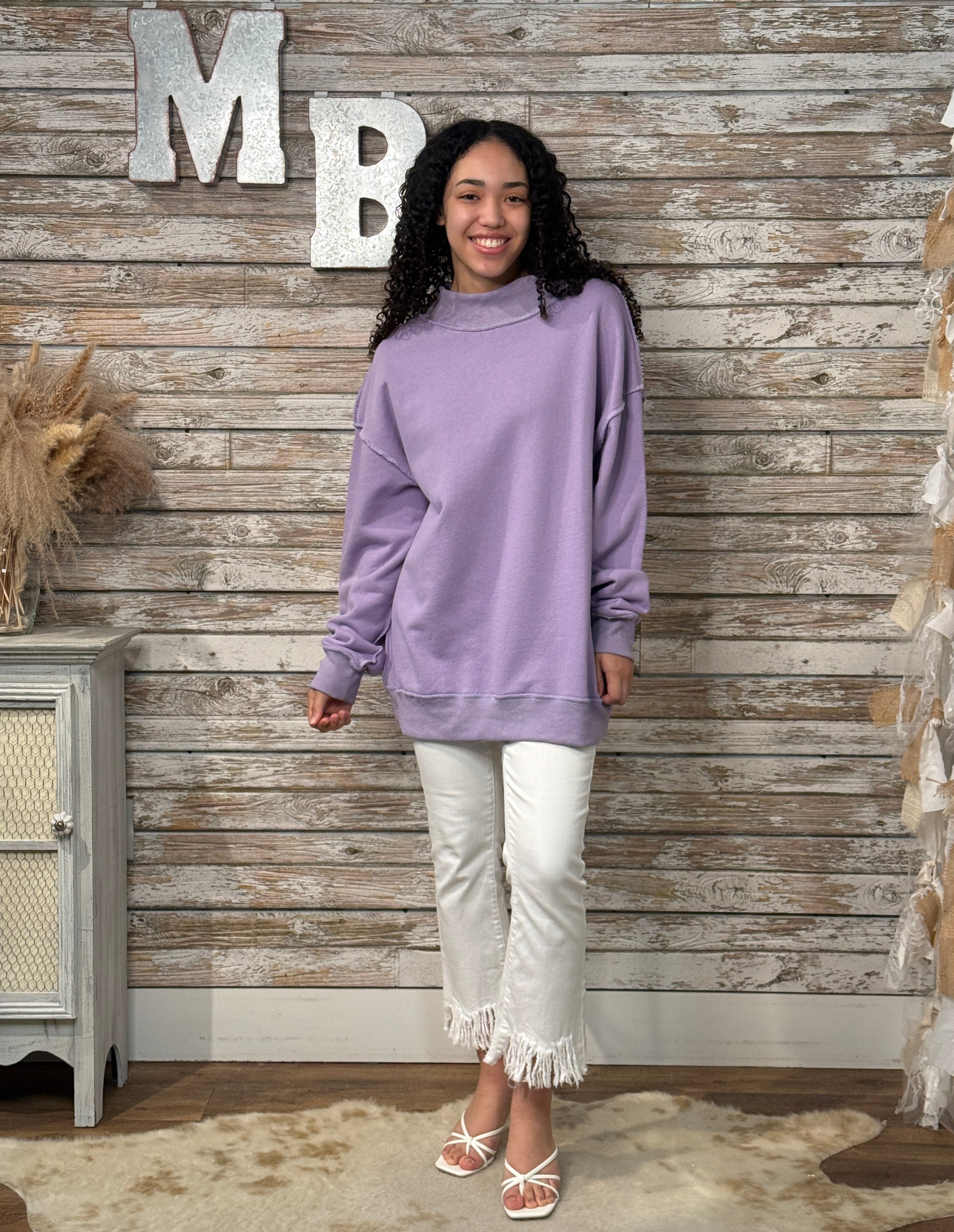 Woman wearing a purple sweatshirt and white pants standing against a wooden wall with decorative letters.
