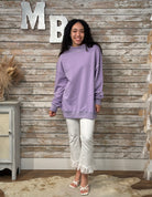Woman wearing a purple sweatshirt and white pants standing against a wooden wall with decorative letters.