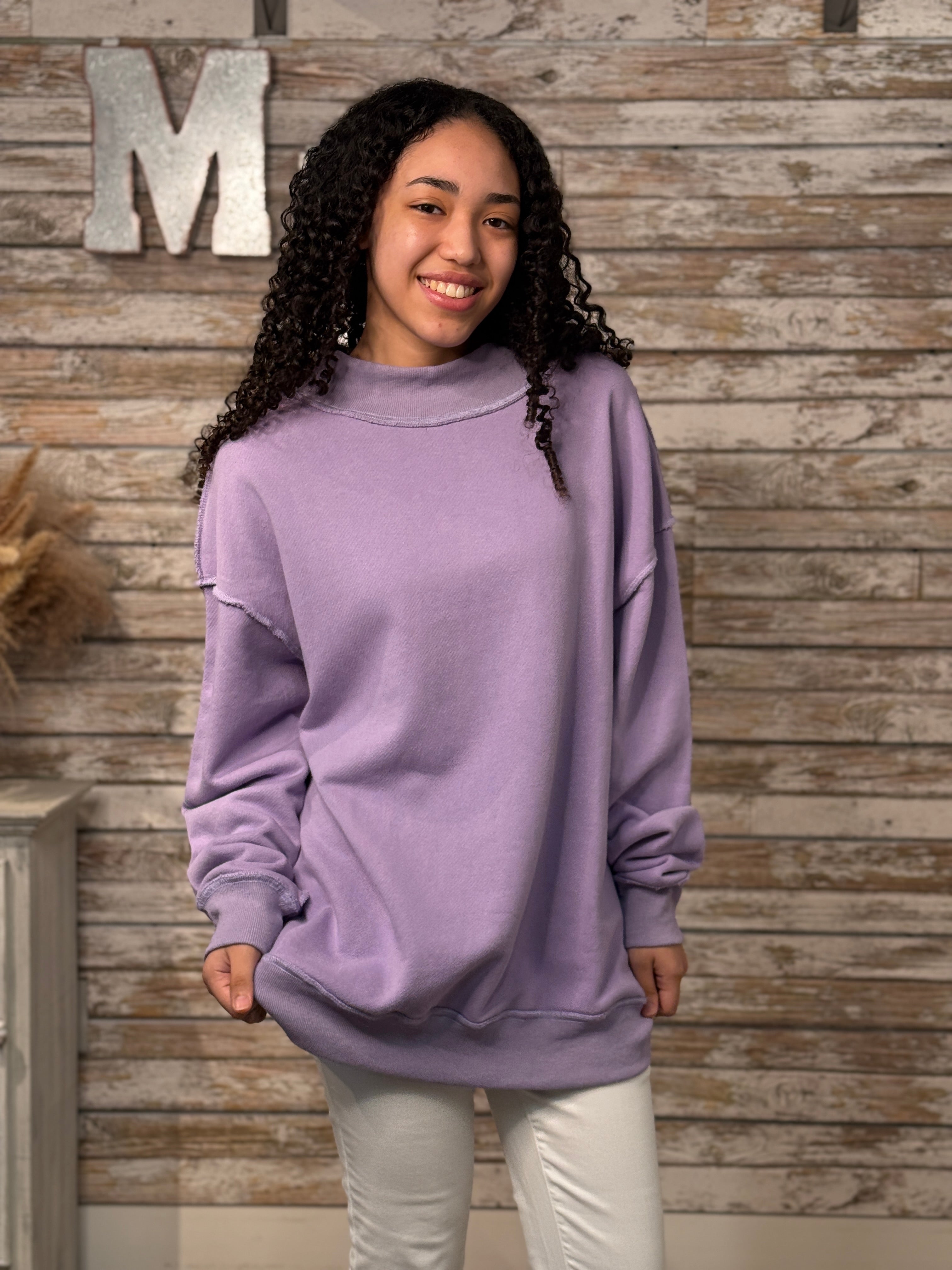 Woman wearing a purple sweater standing against a wooden wall with a decorative 'M' letter.