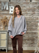 A woman wearing a light gray oversized pullover hoodie with drawstrings and brown pants, standing in front of a rustic wood-paneled wall with decorative letters.