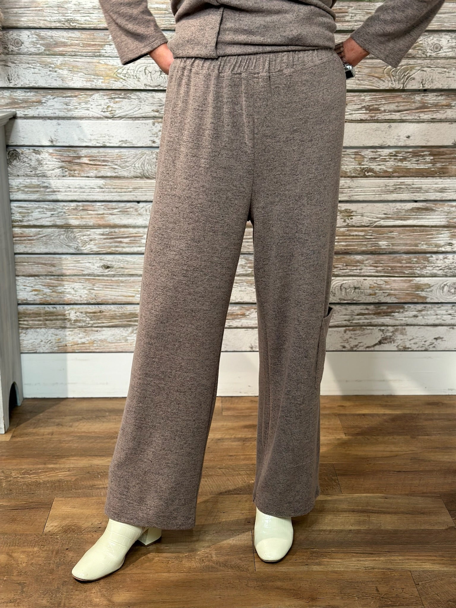A person wearing loose, wide-leg knit pants in a heather taupe-brown color, paired with a matching top and off-white boots, standing against a rustic whitewashed wood plank wall and wooden floor.