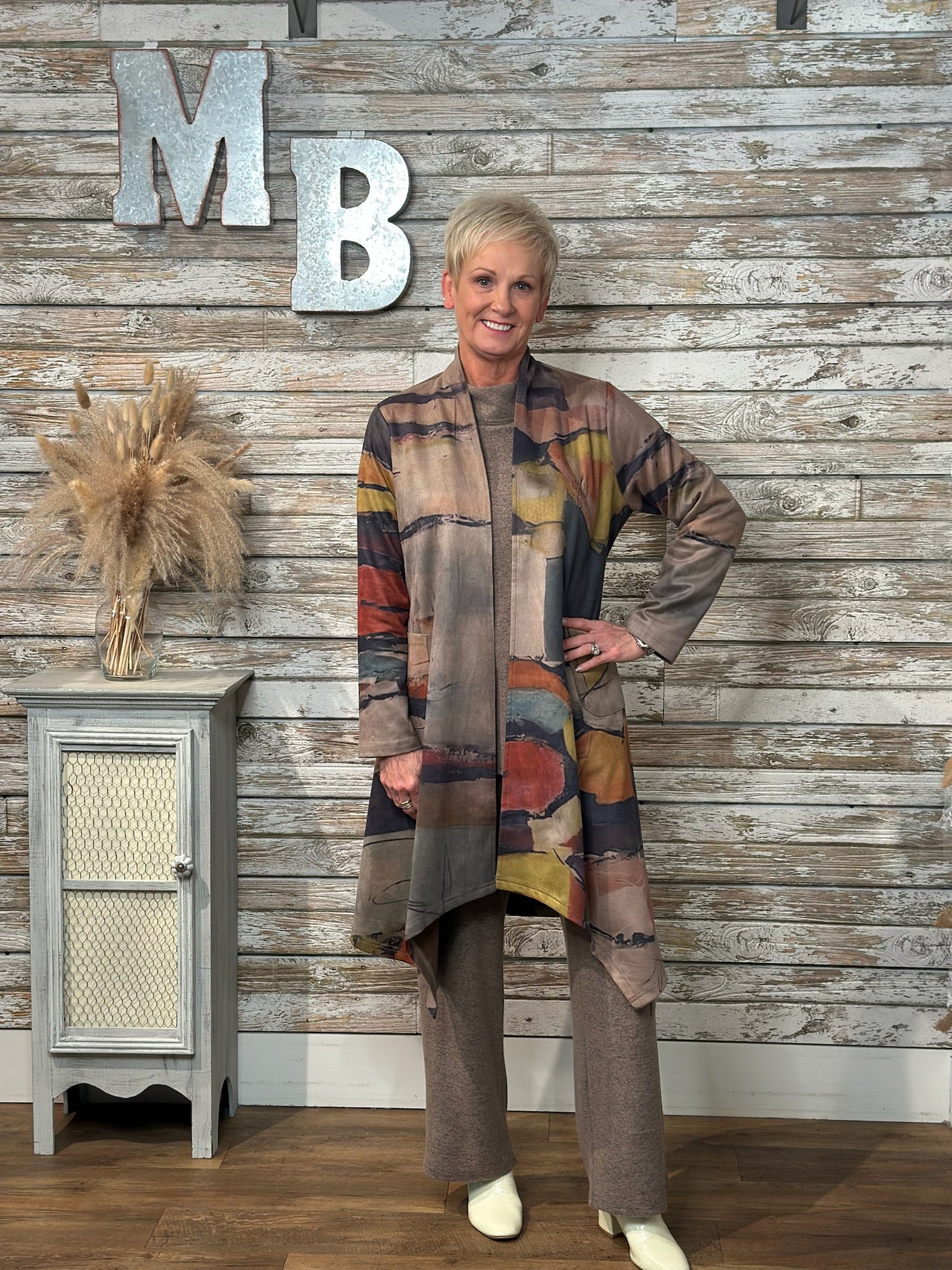 A woman modeling a long, open-front cardigan with an abstract multicolor pattern in earthy tones, paired with matching brown pants, standing in front of a rustic wood-paneled wall with decorative letters.