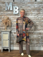 A woman modeling a long, open-front cardigan with an abstract multicolor pattern in earthy tones, paired with matching brown pants, standing in front of a rustic wood-paneled wall with decorative letters.