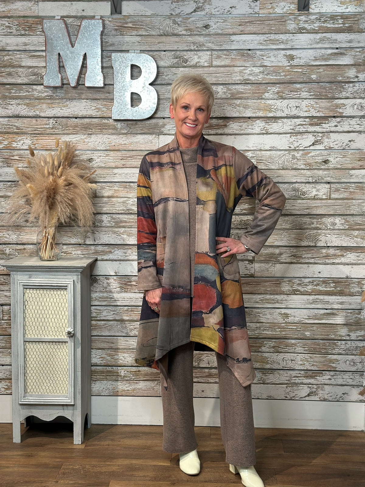 A woman modeling a long, open-front cardigan with an abstract multicolor pattern in earthy tones, paired with matching brown pants, standing in front of a rustic wood-paneled wall with decorative letters.