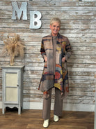 A woman stands in front of a rustic whitewashed wood wall wearing a long abstract-print cardigan over a taupe knit top and matching wide-leg pants, with white boots. Large metal letters 'MB' and a small cabinet with dried grass decor are visible in the background.