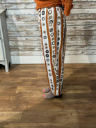 A person wearing loose, wide-leg pants with vertical rust-brown and white stripes, each printed with circular motifs in black, gray, and beige. The person is standing on a wood floor in front of a rustic whitewashed wood wall, wearing a rust-orange top and gray slip-on shoes.