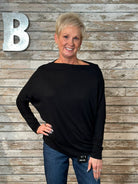 A woman with short blonde hair wearing a black long-sleeve boat-neck top and blue jeans, standing in front of a rustic wood plank wall with a large silver letter 'B' on the left.
