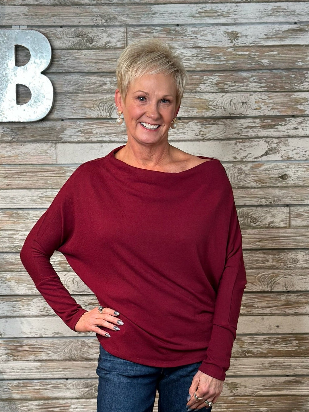 A smiling blonde woman wearing a long-sleeve, boat-neck, slouchy knit top in deep burgundy, paired with blue jeans, standing against a rustic gray wood plank wall with a large silver letter 'B' on the left.