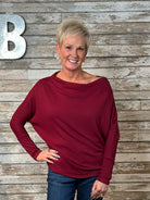 A smiling blonde woman wearing a long-sleeve, boat-neck, slouchy knit top in deep burgundy, paired with blue jeans, standing against a rustic gray wood plank wall with a large silver letter 'B' on the left.
