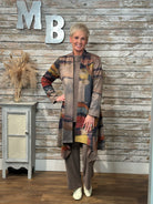 A woman wearing a long patchwork cardigan with abstract multicolor blocks in beige, brown, gray, blue, and rust tones, paired with wide-leg taupe pants and white shoes. 