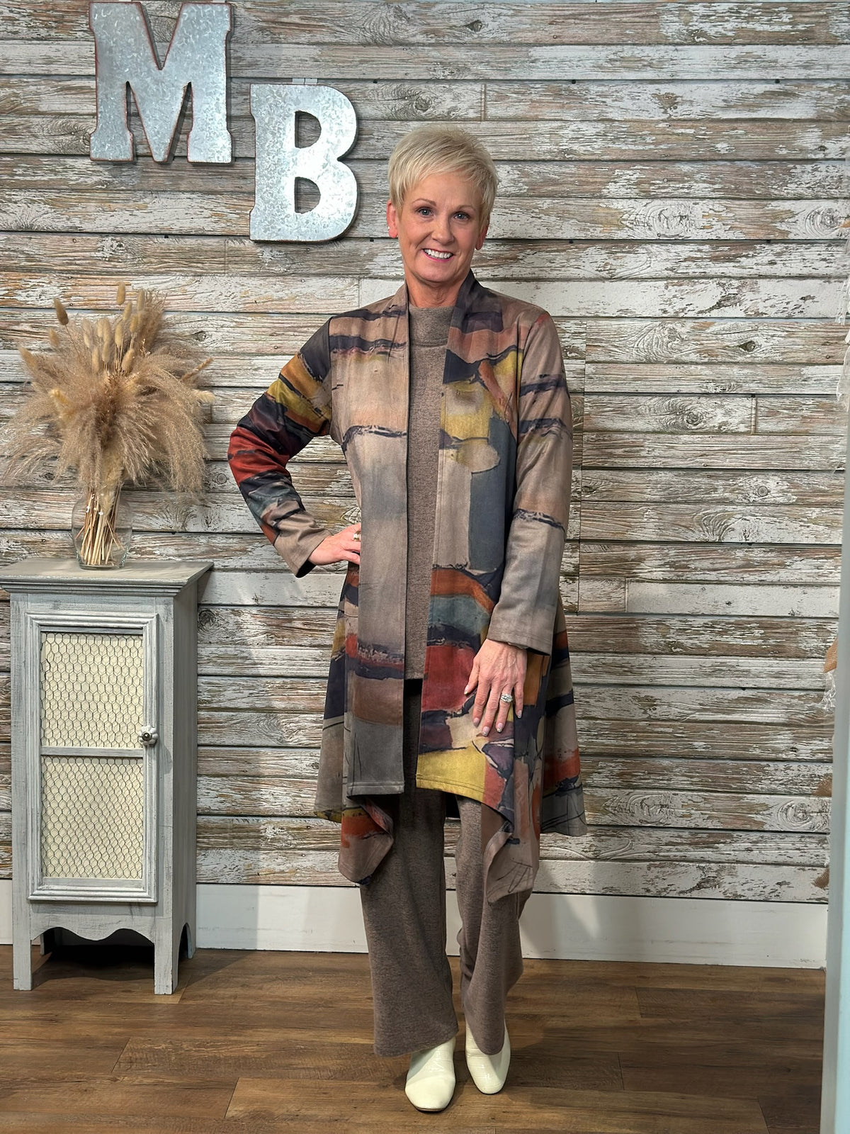 A woman wearing a long patchwork cardigan with abstract multicolor blocks in beige, brown, gray, blue, and rust tones, paired with wide-leg taupe pants and white shoes. 