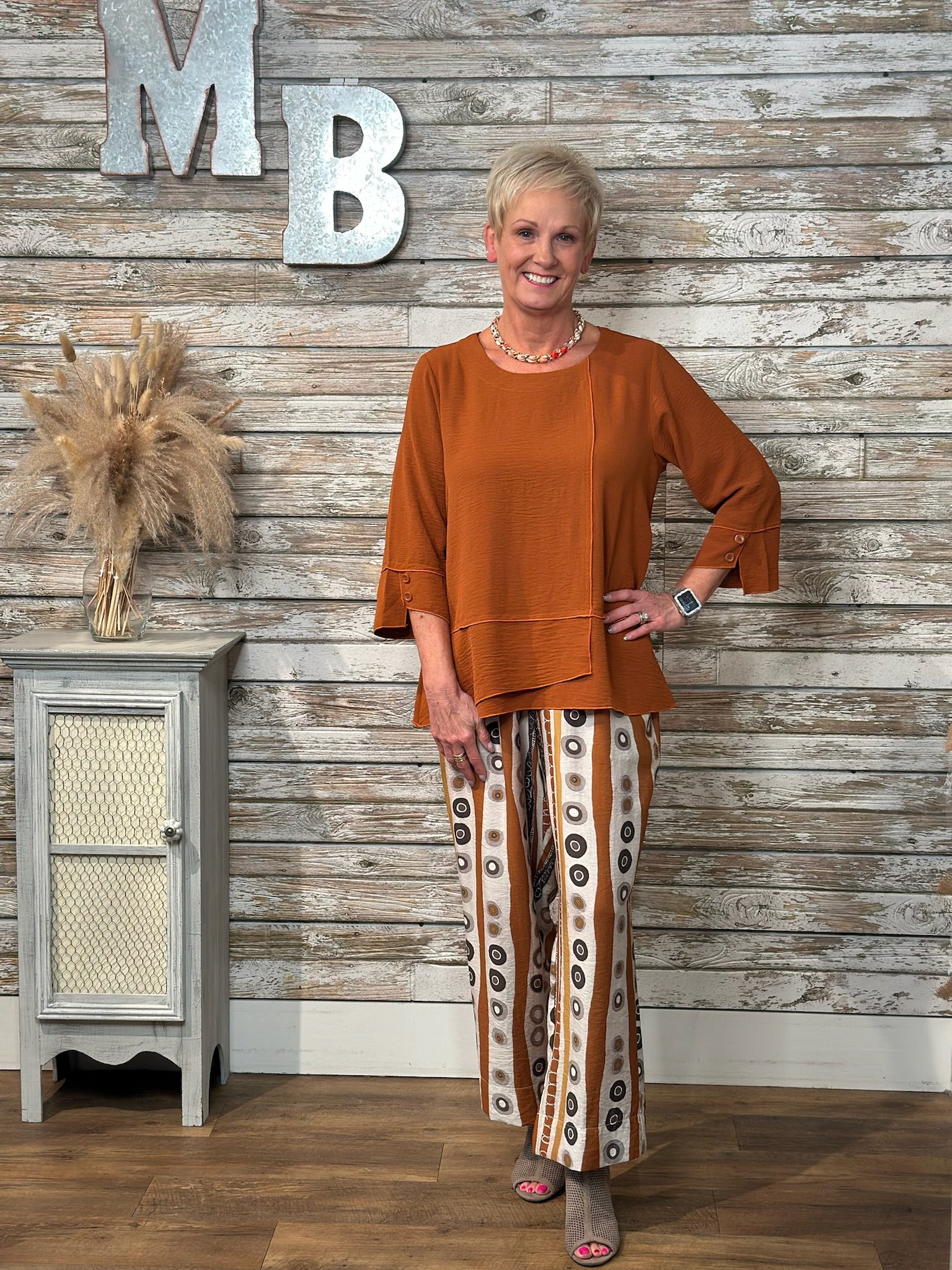 A woman modeling a rust-orange long-sleeve top and patterned wide-leg pants with brown, beige, and white vertical stripes, standing in front of a rustic whitewashed wood wall with decorative letters.