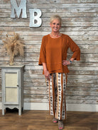 A woman modeling a rust-orange long-sleeve top and patterned wide-leg pants with brown, beige, and white vertical stripes, standing in front of a rustic whitewashed wood wall with decorative letters.