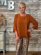 A woman modeling a rust-orange long-sleeve top with patterned pants, standing in front of a rustic wood-paneled wall with decorative letters.