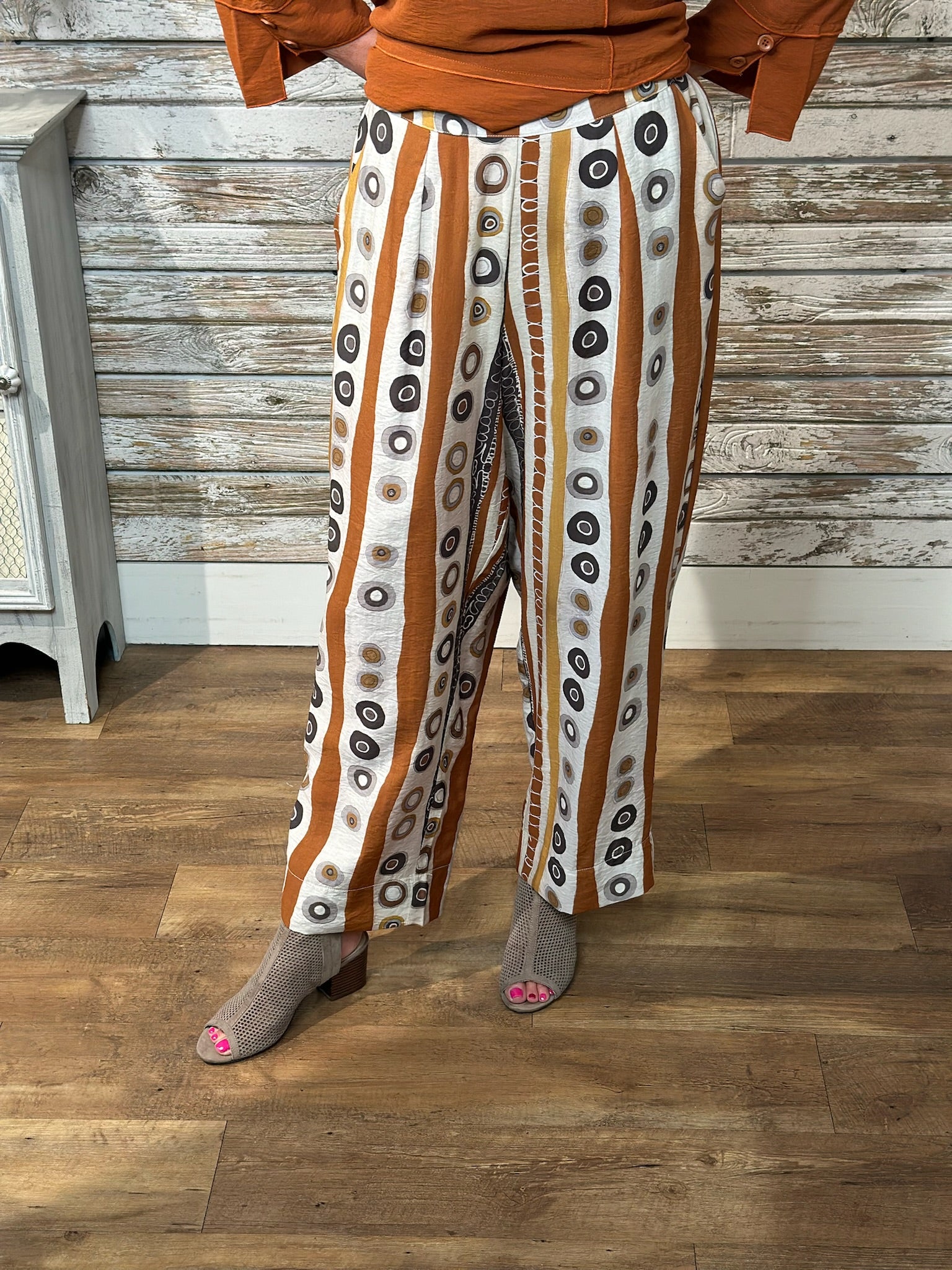 A person wearing wide-leg cropped pants with vertical rust-orange and white stripes and circular black, gray, and brown motifs, paired with an orange top and gray peep-toe heels, standing on a wood floor against a rustic wood plank wall.