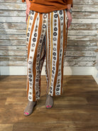 A person wearing wide-leg cropped pants with vertical stripes of brown, white, and black circular motifs, paired with an orange-brown top and gray slip-on shoes, standing against a rustic wood plank wall and floor.