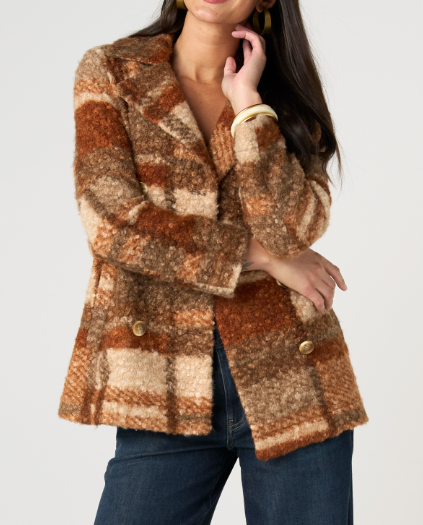 A woman wearing a fuzzy, textured plaid blazer in warm brown, beige, and rust tones, paired with dark blue jeans, against a solid light gray background.