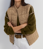 A woman wearing a cropped cable-knit sweater vest in beige over an olive green knit sweater, styled with white pants and a brown belt against a light gray studio background.