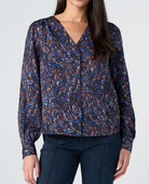 A woman wearing a long-sleeve V-neck blouse with a blue, brown, and white abstract speckled pattern, paired with dark blue jeans, against a light gray studio background.