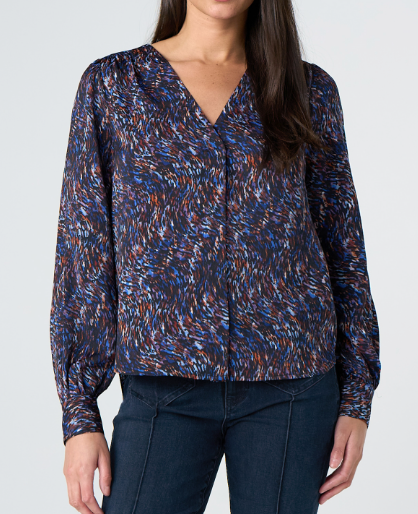 A woman wearing a long-sleeve V-neck blouse with a blue, brown, and white abstract speckled pattern, paired with dark blue jeans, against a light gray studio background.
