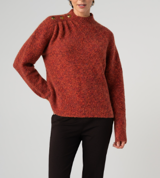 A woman wearing a textured, heathered rust-orange mock-neck sweater with decorative buttons on the shoulder, paired with black pants, against a light gray studio background.