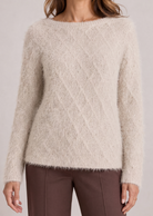 A woman wearing a fuzzy, textured cream-colored knit sweater with a boat neckline and brown trousers, shown from the chest down against a light gray studio background.