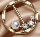 A gold-tone circular brooch or pin featuring a single pearl and three clear rhinestones, displayed on a soft beige satin fabric.