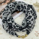 A lightweight scarf with a black leopard print on a light gray background, styled in a loop with gold-toned decorative beads at the end, resting on a floral fabric surface.