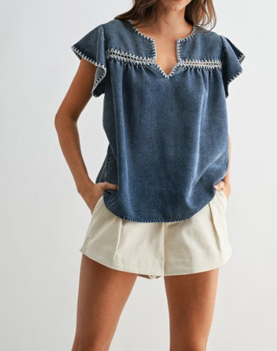 Woman wearing a blue denim top with white trim and beige shorts on a plain background