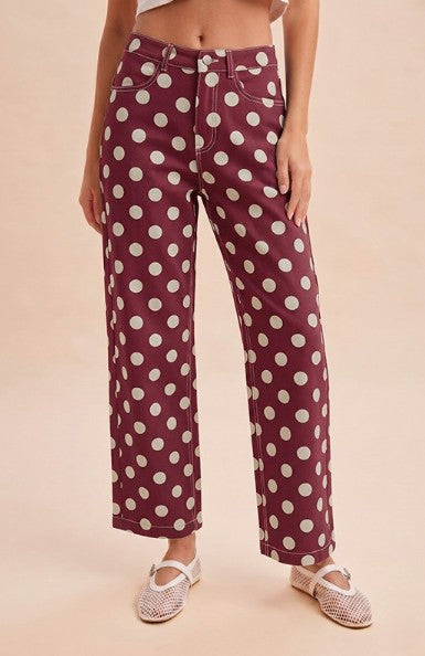 A model wearing high-waisted, wide-leg trousers in a deep burgundy color with a white polka dot pattern, paired with white espadrille flats, set against a solid light beige background.