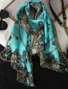 A turquoise scarf with a leopard print border and a black chain-link pattern, styled on a white faux fur surface with sunglasses and a handbag nearby.