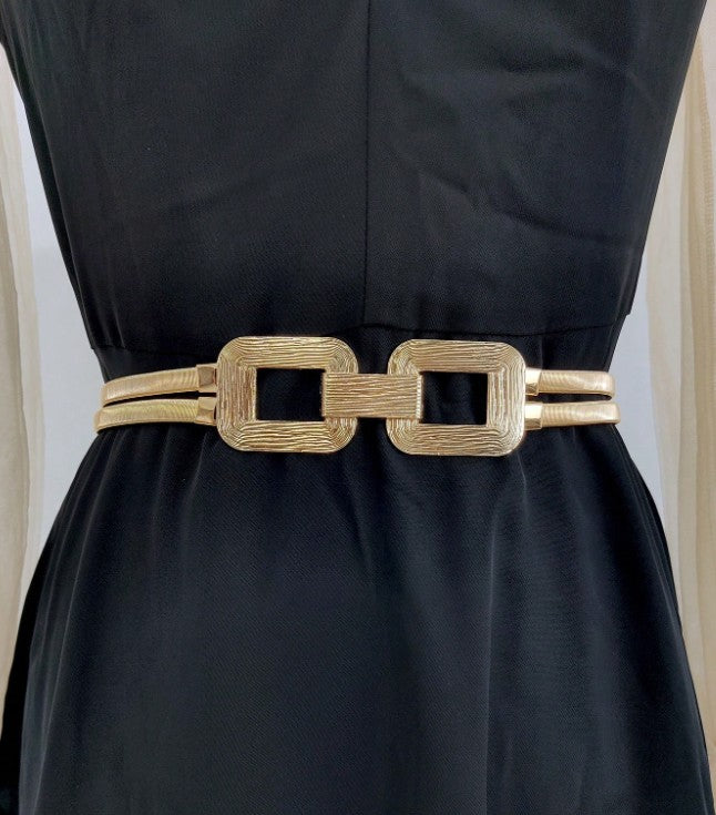 A gold-colored belt with a textured finish and a large rectangular link design, worn as a waist accessory over a black dress.