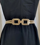 A gold-colored belt with a textured finish and a large rectangular link design, worn as a waist accessory over a black dress.