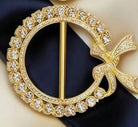 A gold-tone circular brooch with a decorative bow at the right side, set with clear rhinestones, displayed on a dark navy blue fabric background.
