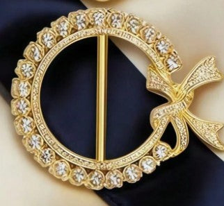 A gold-tone circular brooch with a decorative bow at the right side, set with clear rhinestones, displayed on a dark navy blue fabric background.