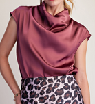 A close-up of a woman wearing a satin-like cowl-neck top in a dusty rose or mauve shade, paired with a leopard-print skirt. The top is the primary product shown, photographed against a plain light background.