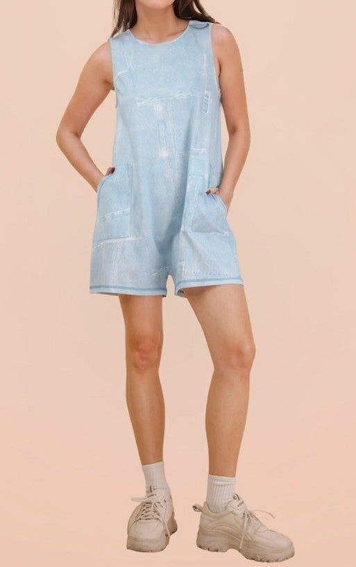A model wearing a light blue denim romper with front patch pockets, paired with white socks and beige sneakers, set against a solid pale peach background.