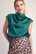 A woman wearing a teal green satin cowl-neck top and a leopard-print skirt, photographed against a plain light beige studio background.